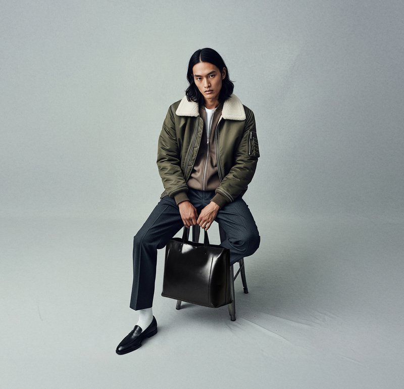 Olive green bomber jacket with a white fleece collar, grey sweater, tailored dark grey trousers, black leather bag, black loafers, and white socks.