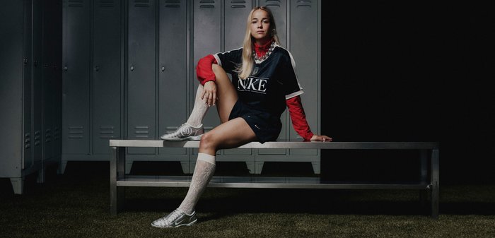 Maglia nera Nike sopra una maglietta rossa a maniche lunghe, abbinata a pantaloni corti neri e scarpe da calcio argentate. Calze al ginocchio bianche a fantasia. Panchina in metallo e armadietti sullo sfondo.