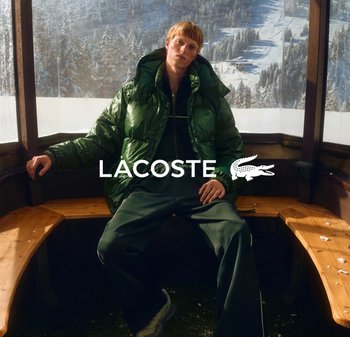 Puffer jacket in shiny green with a hood, worn over a dark green tracksuit. Wooden benches in a snow-covered setting. Brand logo in white.