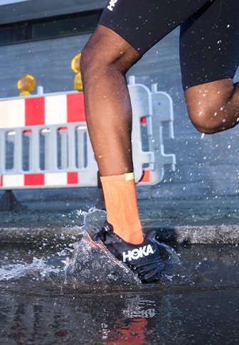 HOKA running shoe in black with distinctive white logo, splashing through water on a wet surface; features breathable material and lightweight design.