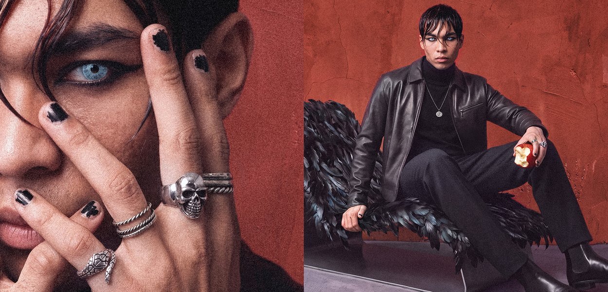 Joven con esmalte de uñas negro y anillos plateados, vistiendo chaqueta de cuero negra y ropa negra, sentado en una silla negra con plumas, sosteniendo una manzana parcialmente comida.