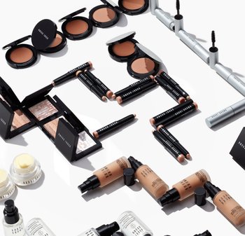 Makeupprodukter arrangerade på en vit bakgrund, inklusive svarta kompaktförpackningar, krämiga pennor och olika flaskformer i nude- och genomskinliga färger.