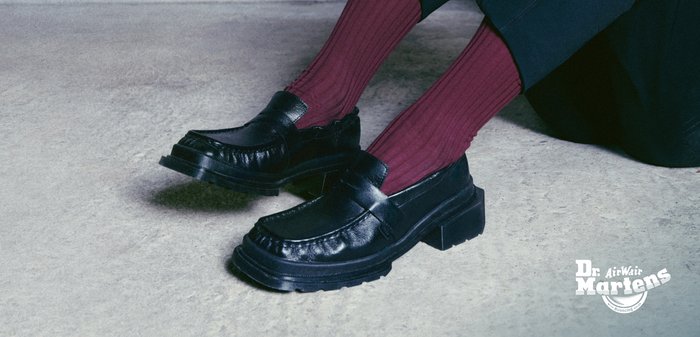 Mocasines de cuero negro con una suela gruesa, que presentan costuras decorativas y una cinta cruzando el empeine, combinados con calcetines burdeos acanalados.