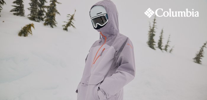 Hellviolette Skijacke mit orangefarbenen Akzenten, ausgestattet mit Kapuze, Reißverschlusstasche und lockerem Schnitt. Trägt eine Skimaske und Skibrille.