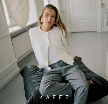 Weißer, gestrickter Cardigan mit dekorativen Bändeln, darüber ein weißes Top. Graue, maßgeschneiderte Hosen und schwarze Schuhe, sitzend auf einem strukturierten schwarzen Kissen.