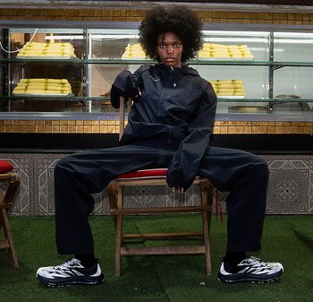 Veste coupe-vent noire et pantalon à coupe oversized, associés à des baskets blanches et noires, sur un fond texturé avec des carreaux décoratifs.