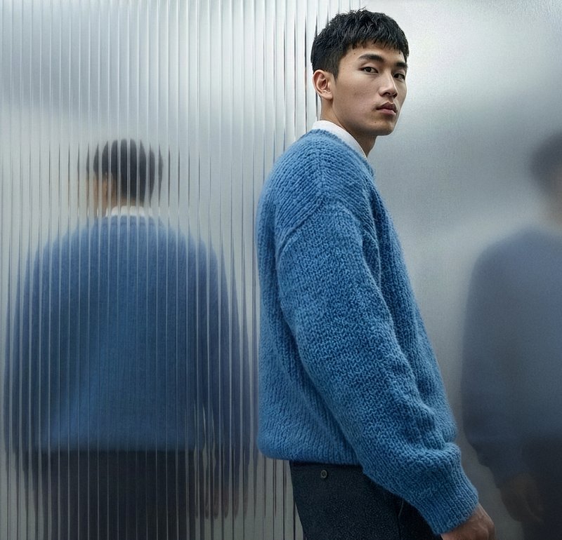Νεαρός άνδρας με μπλε πλεκτό πουλόβερ στέκεται δίπλα σε έναν τοίχο από υαλοπίνακα με υφή, που αντανακλά δύο θολές εικόνες του.