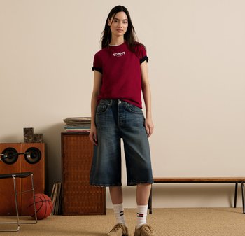 T-shirt bordeaux avec le logo "TOMMY", manches retroussées ; culottes en denim bleu ; baskets beige avec des accents blancs et rouges ; arrière-plan assis avec des textures en bois.