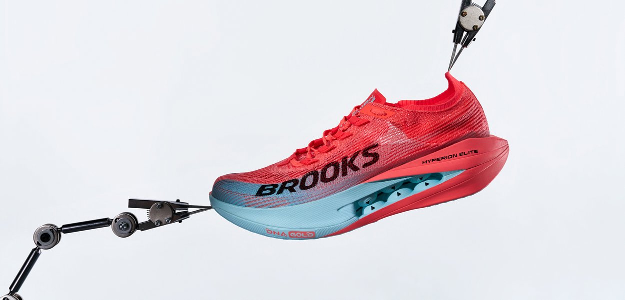 Zapatilla de correr Brooks Hyperion Elite en rojo y turquesa sostenida por brazos robóticos sobre un fondo blanco, mostrando los detalles de la suela y la marca con texto.