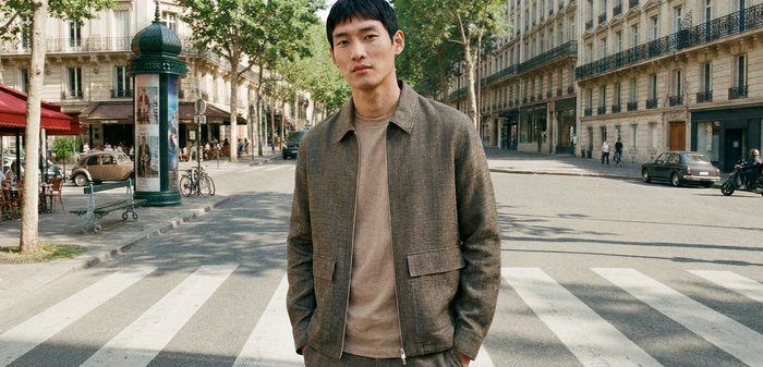 Mladý muž v hnedej bunde a nohaviciach stojí na mestskom priechode pre chodcov s stromami a historickými budovami v pozadí.