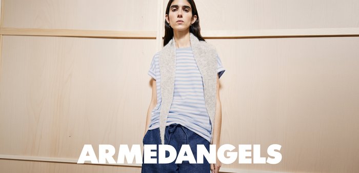 Junge Frau trägt ein gestreiftes T-Shirt, blaue Hose und eine graue ärmellose Weste, steht vor einer hellen Holzwand mit dem Text "ARMEDANGELS".