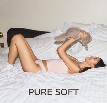 Vrouw in lichtroze lingerie liggend op een wit bed, glimlachend en een pluizige grijze kat boven haar hoofd houdend. Tekst: "PURE SOFT."