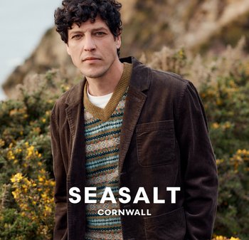 Hnědá kordová bunda přes svetr s multikolorovým vzorem a výstřihem do V. Pozadí sází na zeleň a skály, což naznačuje venkovní prostředí.