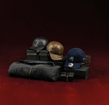 Trois casquettes en noir, marron et bleu marine posées sur des boîtes noires avec le logo New Era, à côté d'une veste zippée New Era gris foncé pliée sur une surface rouge.