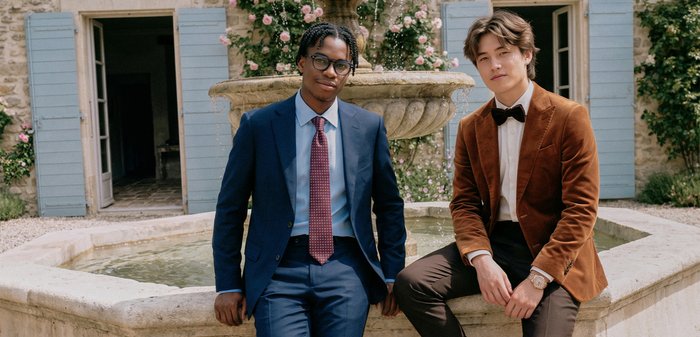 Deux hommes en tenue formelle, l'un en costume bleu avec cravate rouge, l'autre en blazer marron et nœud papillon, assis près d'une fontaine en pierre en plein air.