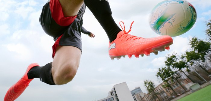 Un joueur de football portant des crampons rouges saute, se préparant à frapper un ballon blanc avec des marques bleues et rouges sur un terrain extérieur.