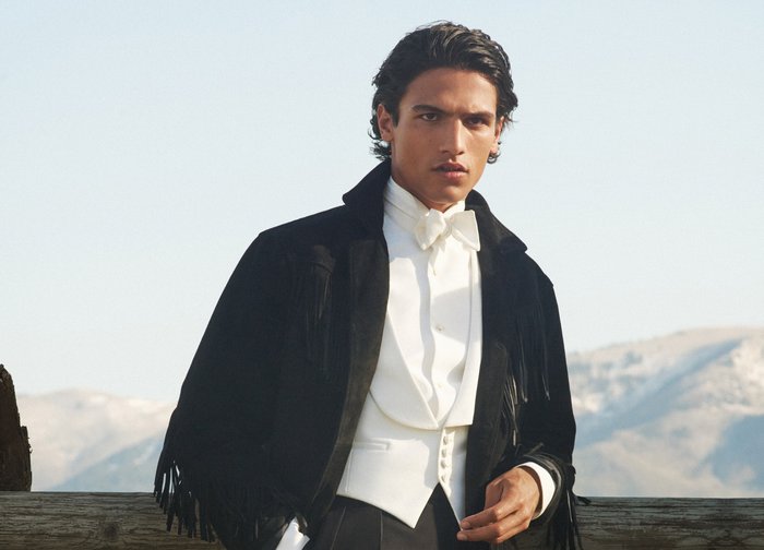 Ung man klädd i en vit skjorta med fluga och svart fransjacka, står utomhus med berg i oskärpa i bakgrunden.