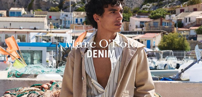 Giovane uomo con camicia a righe aperta e giacca beige in piedi accanto a reti da pesca e barche in un porto soleggiato con edifici dai colori pastello sullo sfondo.