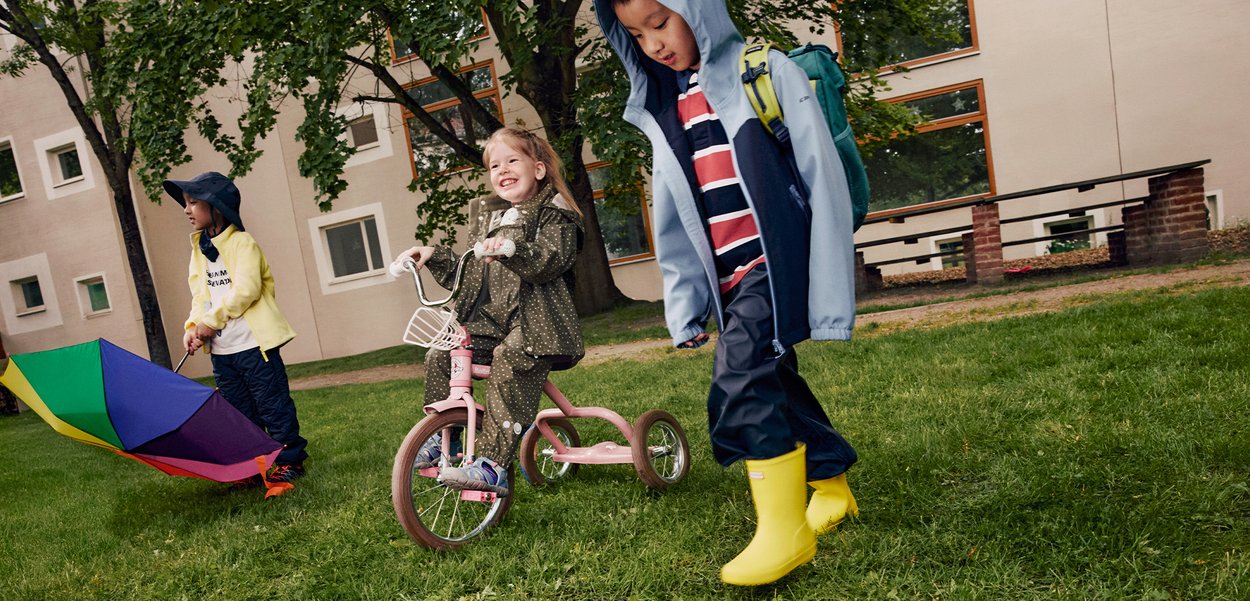 Children outdoors: one rides a pink tricycle with a white basket; others hold colourful umbrellas and wear waterproof clothing and yellow boots.