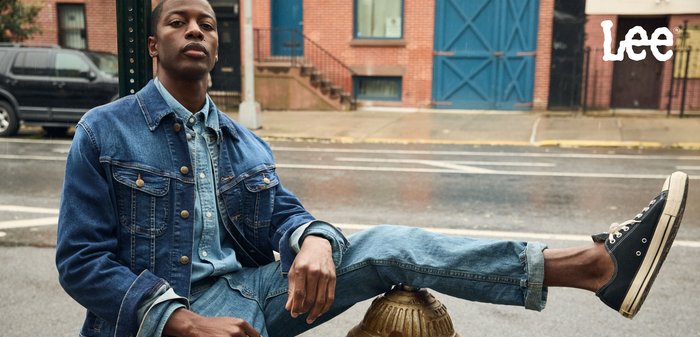 Jeansjacka och ljusblå skjorta med jeans. Svarta sneakers, sittande på en brandpost. Urban miljö med tegelbyggnader i bakgrunden.