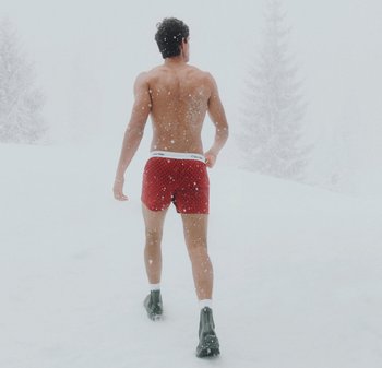 Man i röda shorts och svarta stövlar går barbröstad genom ett snötäckt landskap med snö som faller runt honom.