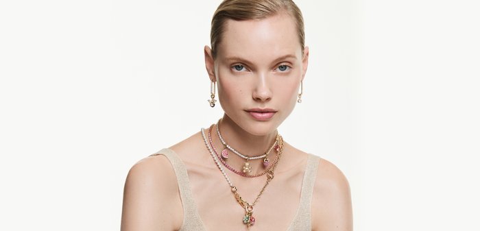 Woman wearing layered gold and pink gemstone necklaces and gold drop earrings with pink and clear stones against a white background.