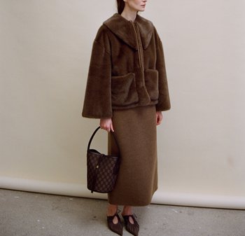 Brun faux päls zip-up jacka med en stor krage, kombinerad med en lång brun kjol. Tillbehör inkluderar en mönstrad handväska och bruna skor.