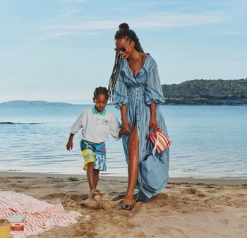 Donna in abito a righe tiene la mano di un ragazzo mentre camminano a piedi nudi su una spiaggia sabbiosa vicino a un mare calmo sotto un cielo sereno.