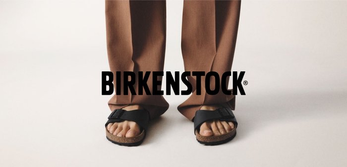 Füße, die schwarze Birkenstock-Sandalen tragen und auf einer hellen Oberfläche stehen, mit braunen Hosen, die die Knöchel bedecken.