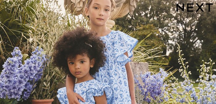 Due ragazze giovani in abiti floreali blu coordinati stanno vicine in un giardino con grandi vasi di terracotta e fiori viola.