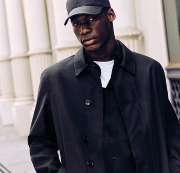 Ung man som bär svart keps, svart knäppt kappa över en svart skjorta och vit t-shirt, stående vid vita klassiska kolonner.