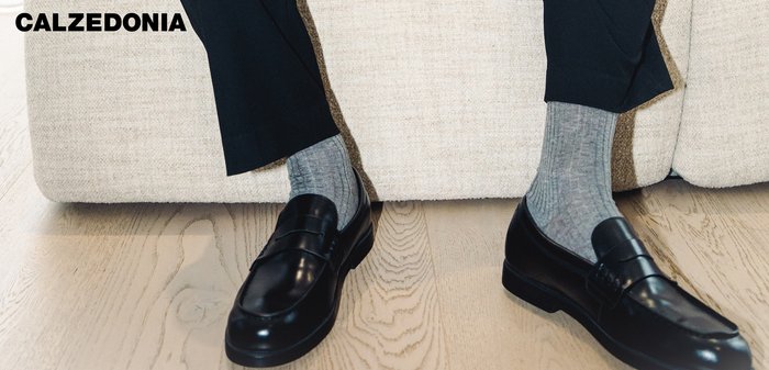 Person sitting on beige sofa wearing black loafers, gray socks, and black pants on light wood floor; "CALZEDONIA" text in top left.