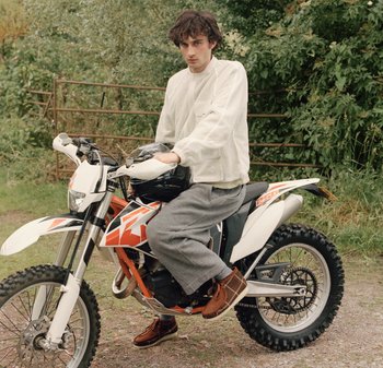 Veste blanche texturée, pantalon de survêtement gris et chaussures brunes portés par une personne sur une moto blanche avec des accents rouges, dans un cadre verdoyant.