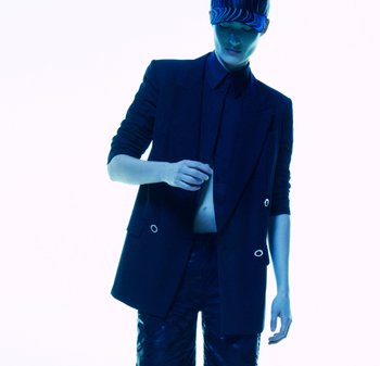 Niebieski oversize'owy blazer z srebrnymi detalami i ciemną koszulą pod spodem. Pasujące spodnie w wzory. Model nosi czapkę w paski. Białe tło.