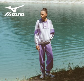Sudadera deportiva Mizuno en gris claro y morado con capucha, logo en el pecho. Pantalones de chándal morados a juego con puños elásticos. Agua al fondo.