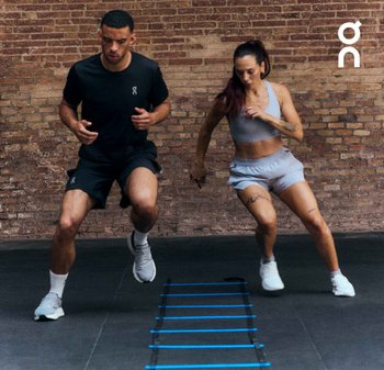 Twee individuen voeren agility-oefeningen uit op een gymvloer, gekleed in zwarte en lichtgrijze sportoutfits. Een blauwe agility ladder is zichtbaar.