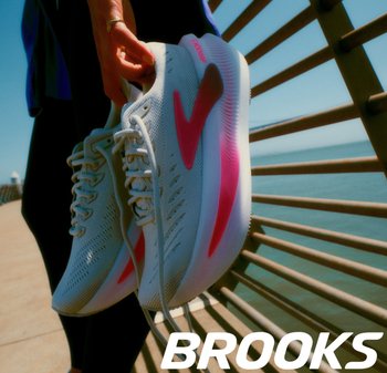 Witte hardloopschoenen van Brooks met roze accenten, ademend bovenwerk van mesh en een gestructureerde zool, gehouden tegen een achtergrond van water en een reling.