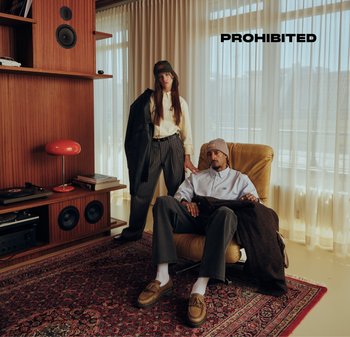 Two individuals stand in a cozy, modern interior. The woman wears a long-sleeve shirt and striped pants, while the man is in a light jacket and beanie. A retro wooden shelf with speakers and a red lamp is in the background.