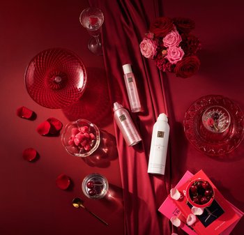 Tre kosmetikkflasker (rosa og hvite) på en rød stoffbakgrunn, omgitt av en rød glassbolle med bringebær og et blomsterarrangement.