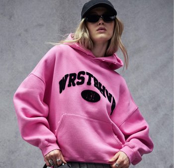 Persona che indossa una felpa oversize di un rosa acceso con testo nero, cappello nero, occhiali da sole scuri e anelli, posando davanti a uno sfondo grigio uniforme.