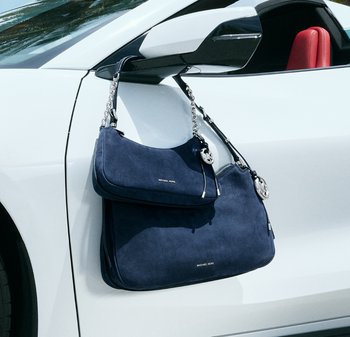 Dos bolsos Michael Kors de color azul marino colgando del espejo lateral de un coche blanco, con el interior rojo visible a través de la ventana.