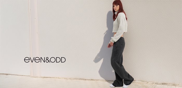 Verkorte, lichtgrijze gebreide trui gecombineerd met losse zwarte jeans en witte sneakers, tegen een gestructureerde lichte achtergrond.