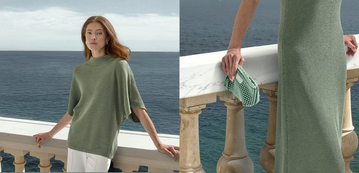 Frau trägt ein salbeigrünes Strickoberteil und Kleid, steht an einem steinernen Balustradenpfosten mit Blick auf das Meer und hält eine grün-weiße Clutch.