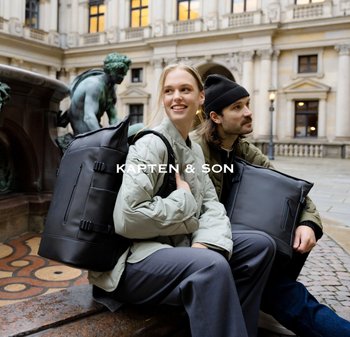 Dva lidé sedí venku u kamenné fontány, každý nese velký černý batoh, s historickou budovou v pozadí.