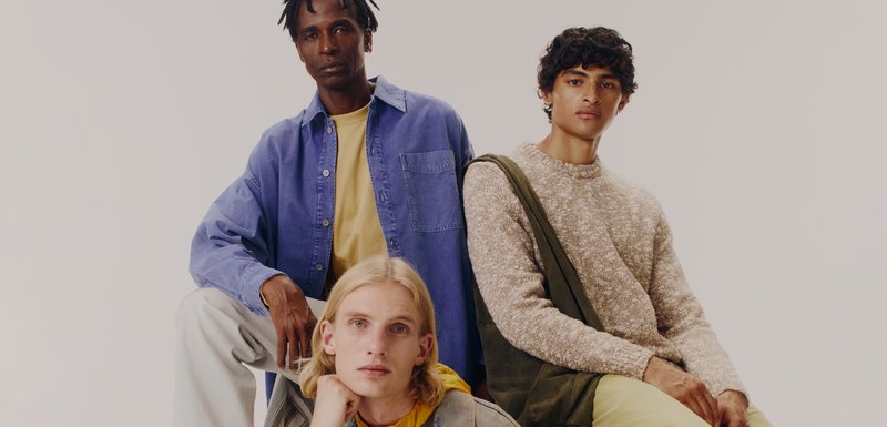 Trois individus posent ensemble. Celui de gauche porte une chemise bleue à boutons, celui du centre un hoodie jaune, et celui de droite un pull en tricot beige avec des accents verts.