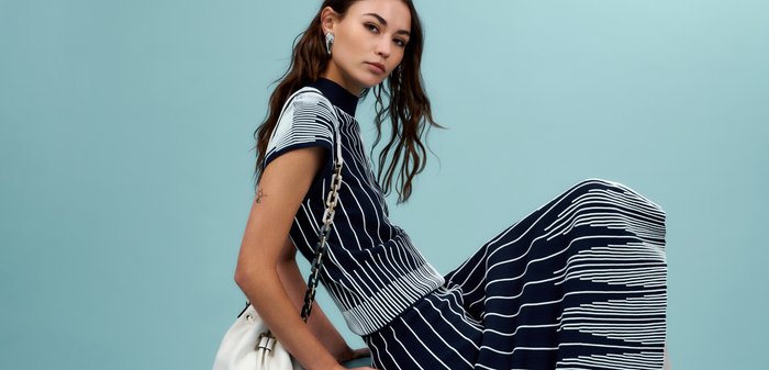 Kvinde sidder på tværs på en skammel iført en sort og hvid stribet kjole, hvide støvler og holder en hvid håndtaske mod en blå baggrund.