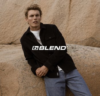 Hemd aus schwarzem Cord mit Knopfleiste, kombiniert mit einem weißen T-Shirt und blauen Jeans. Das Model posiert auf einer felsigen Oberfläche, das Logo ist gut sichtbar.