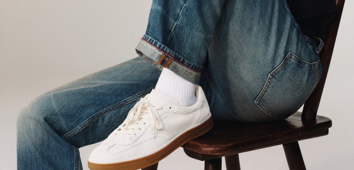 Személy egy fazsámolyon ül, felhajtott kék farmert, fehér zoknit és fehér cipőt visel gumitalppal.