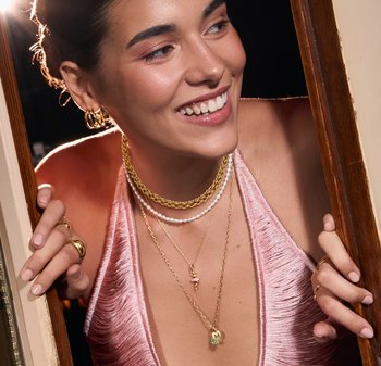 Collane a strati in oro e argento, dettagli in perle e orecchini a cerchio indossati con un abito rosa cipria testurizzato, che mettono in risalto i dettagli dei gioielli preziosi.