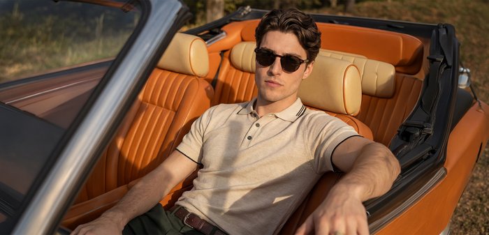 Junger Mann mit Sonnenbrille und beige Poloshirt, sitzt im orangefarbenen Leder-Cabriolet-Innenraum im Sonnenlicht.